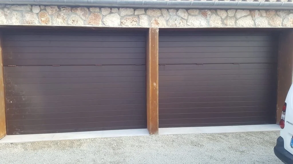 Dos puertas de garaje de color marrón, con estructura de madera y muro de piedra en la parte superior, en un entorno exterior.