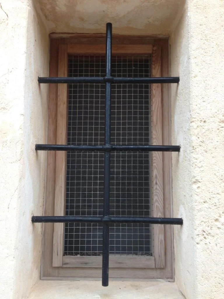 Ventana con barrotes de hierro y malla metálica, rodeada de marco de madera, en una pared de color beige.