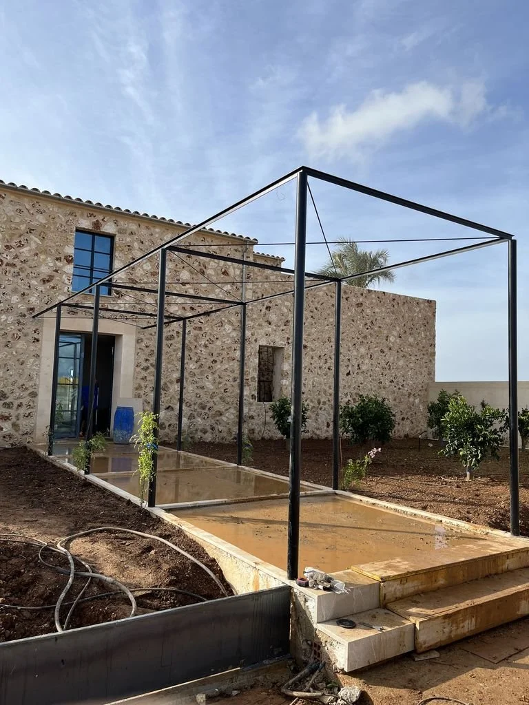Estructura metálica en una terraza en construcción, con tierra y plantas pequeñas alrededor, y un edificio de piedra al fondo, bajo un cielo despejado.