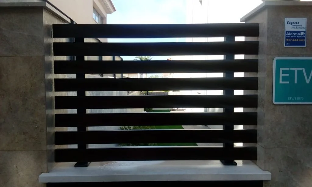 Puerta de reja negra con marco de metal en la entrada de una propiedad, con señales de alarma y CCTV a la derecha.