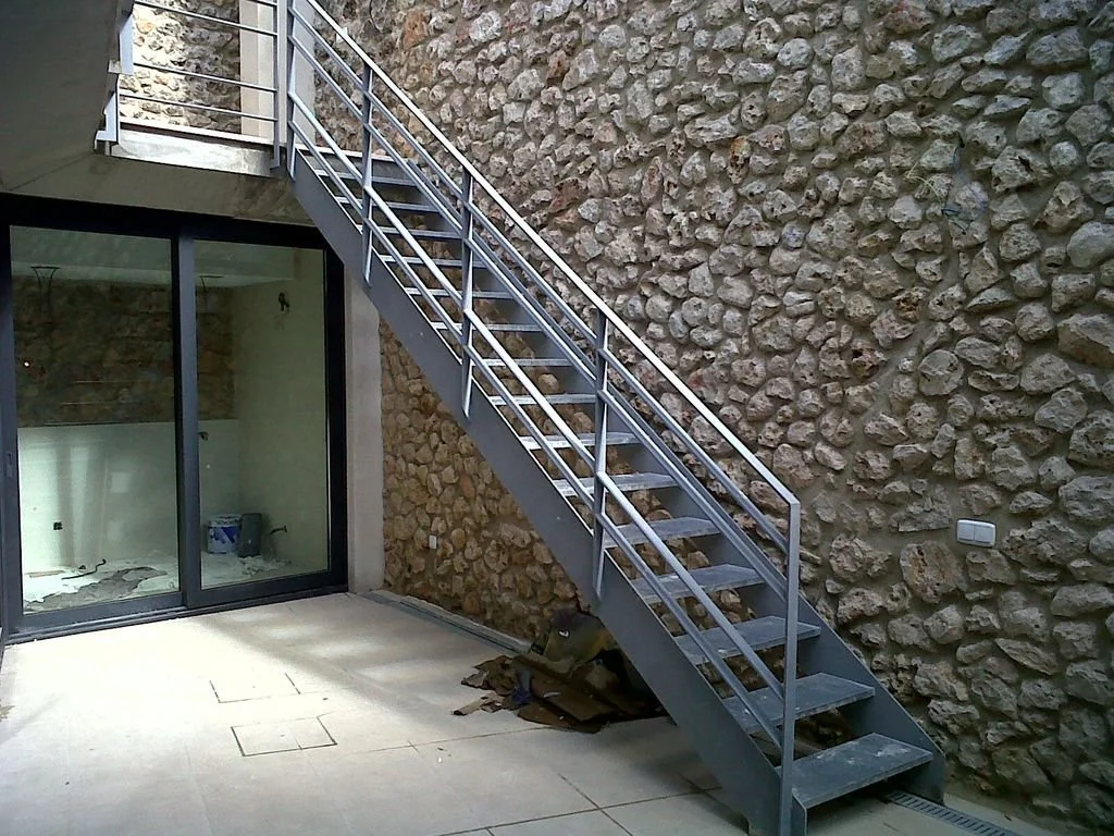 Escalera de metal en un patio con muro de piedras y ventana de vidrio con marco negro.