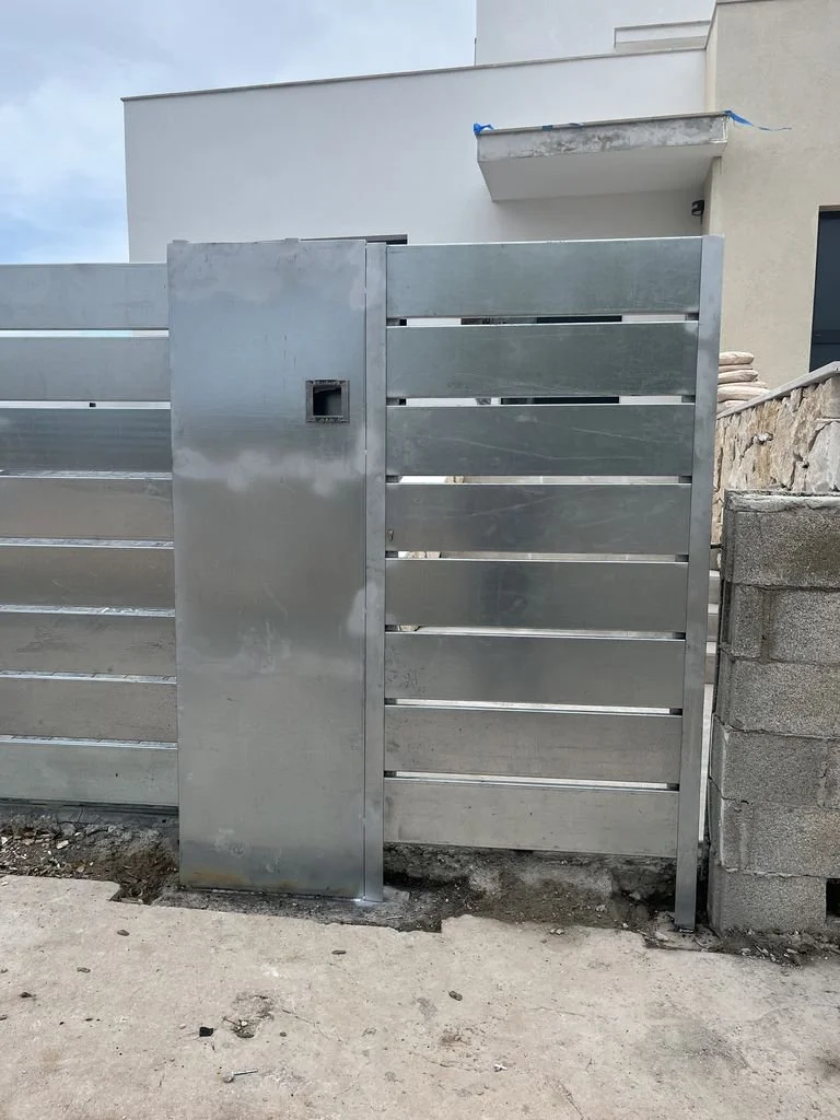 Puerta metálica de reja instalada en una entrada con muro de bloques de concreto del lado derecho y pared de edificio al fondo.