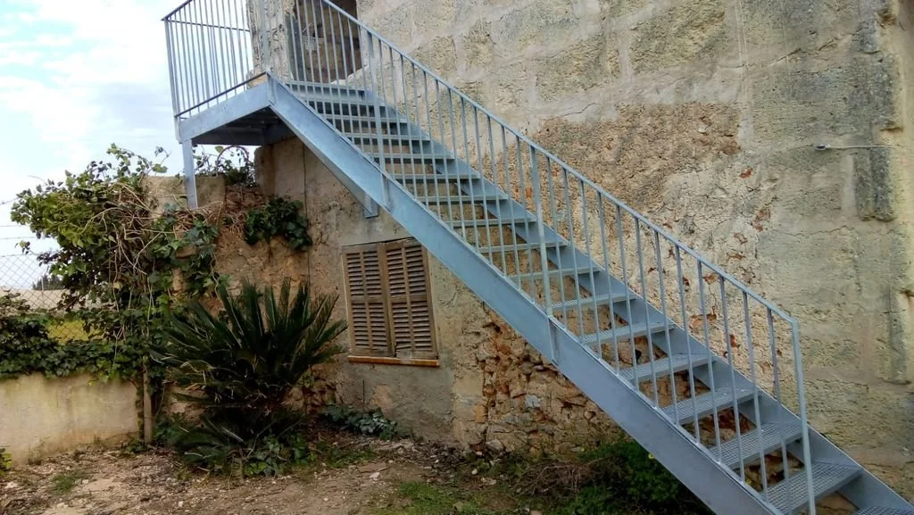 Escalera de metal azul que conecta el suelo con un balcón en una pared de piedra, frente a una planta de palmera y enrejado lateral en un área exterior.