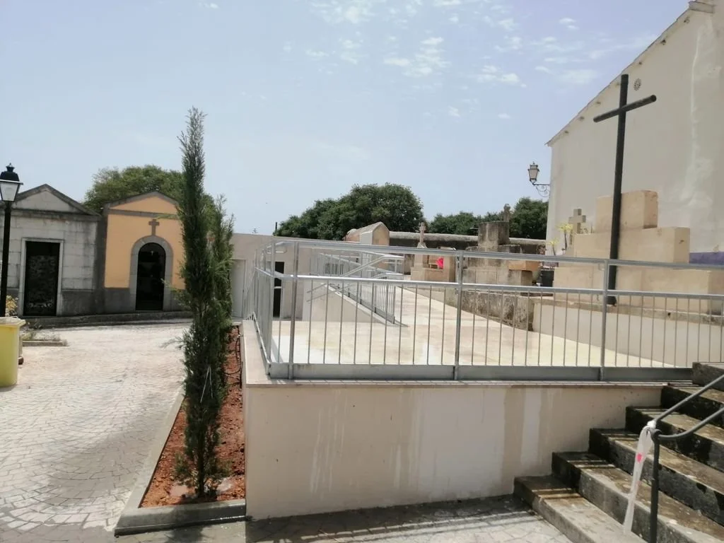 Patio con camino de piedra, cercado de barandillas metálicas, escaleras de piedra, plantas y cruces en las paredes de edificios cercanos.