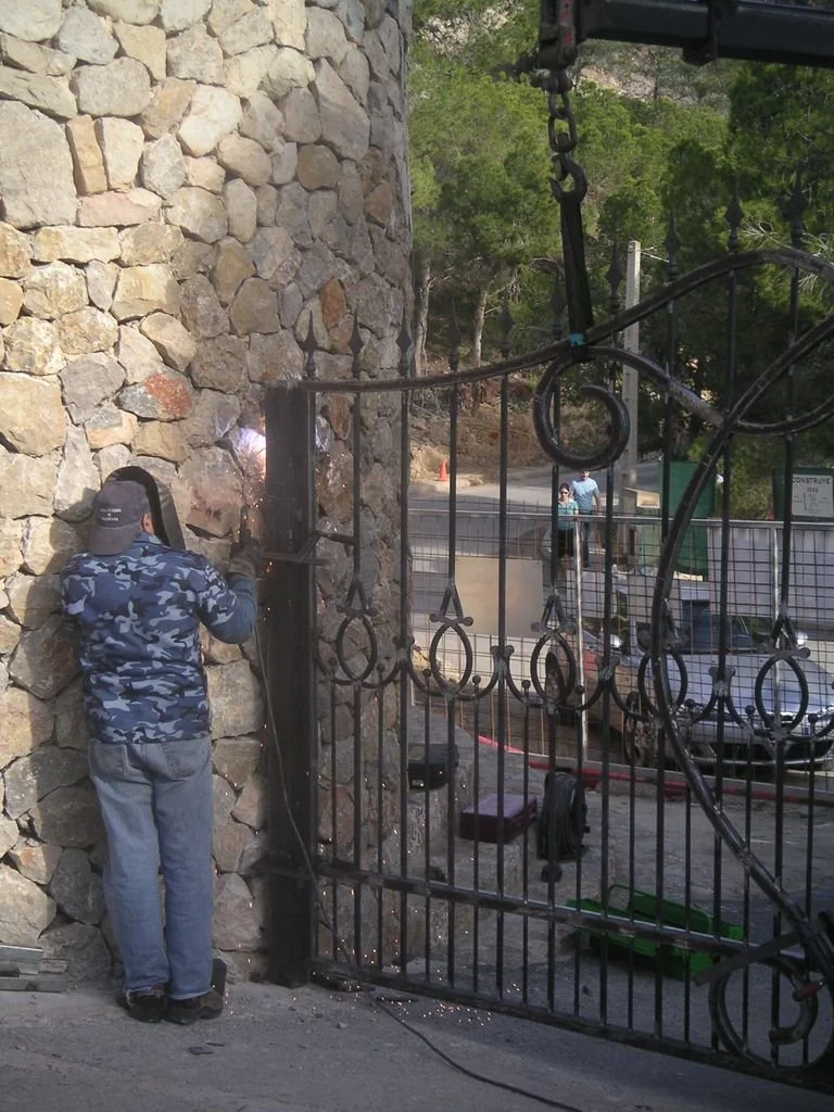 Persona soldando una reja de metal junto a una pared de piedra, en un área exterior.