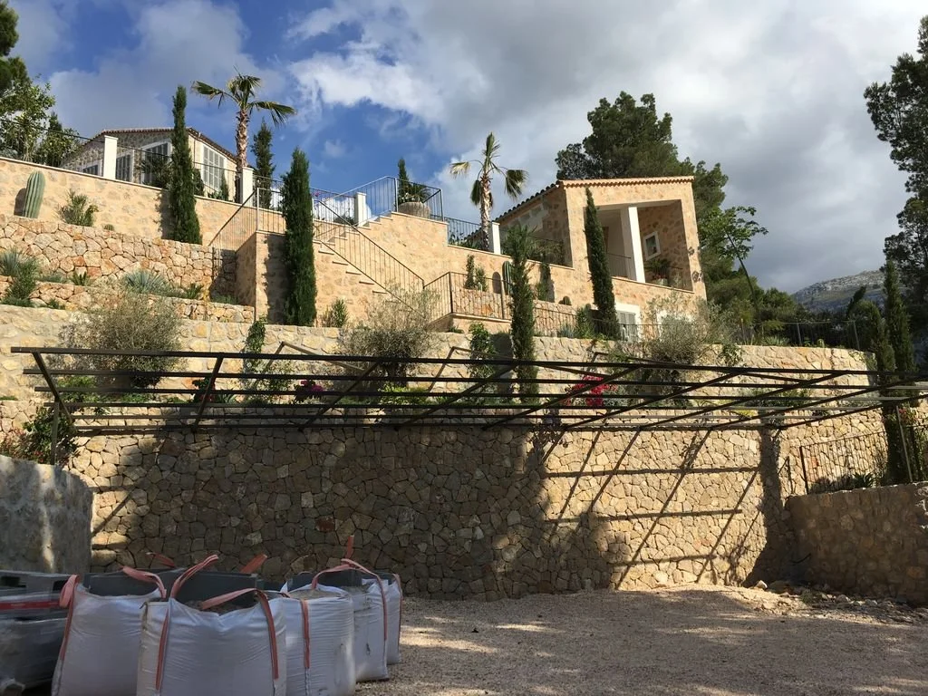 Vista de una casa en una colina con paredes de piedra, árboles y plantas, incluyendo palmeras y cipreses, con sombra en primer plano y un cielo nublado.