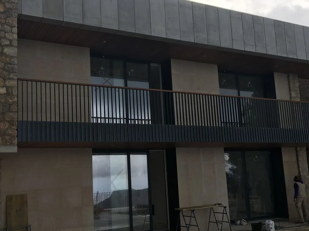 Vista de la fachada de un edificio moderno con paredes de piedra y ventanas grandes con vidrios oscuros. Hay un balcón con barandal de metal y una persona en el extremo derecho.