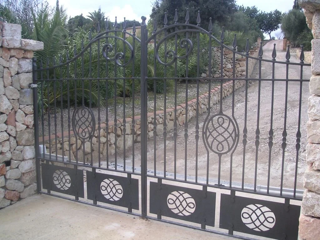 Puerta de reja negra con diseño ornamental, entre muros de piedra, con camino de tierra y vegetación al fondo.