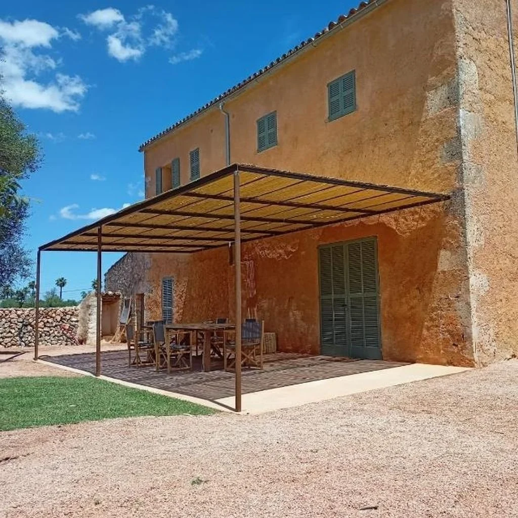 Vista de una casa con paredes de color ocre, ventanas con persianas grises, y un área de descanso exterior con mesa y sillas de madera bajo un toldo de color beige. Al fondo, un muro de piedras y árboles.