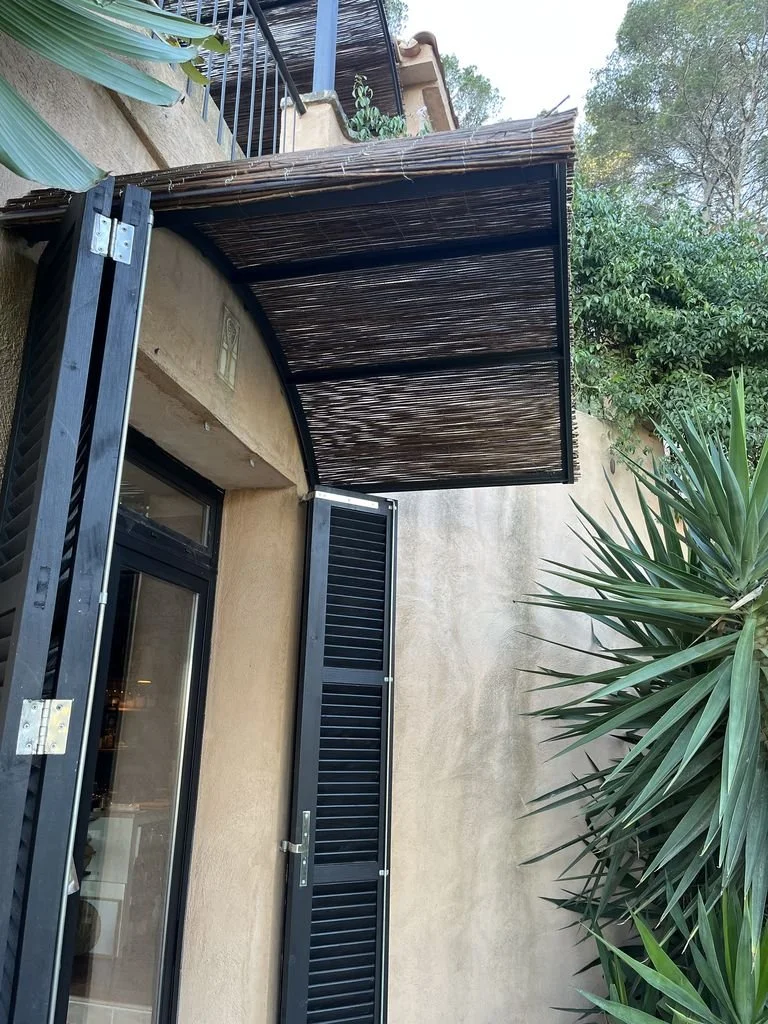 Mini pérgola de madera y metal sobre una puerta de cristal con persianas negras, rodeada de plantas verdes y árboles.