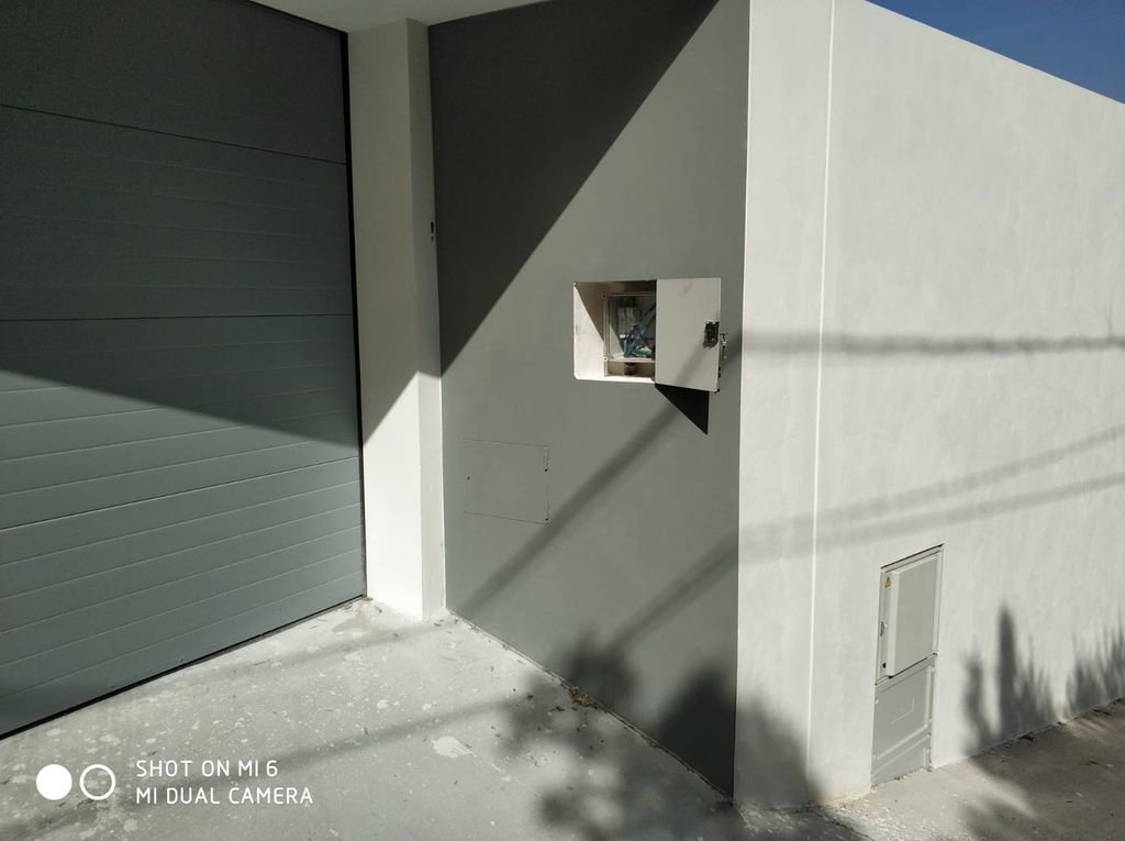 Fachada de una construcción moderna con una puerta de garaje gris y una pared gris, sombra proyectada y caja de conexiones eléctricas abiertas.