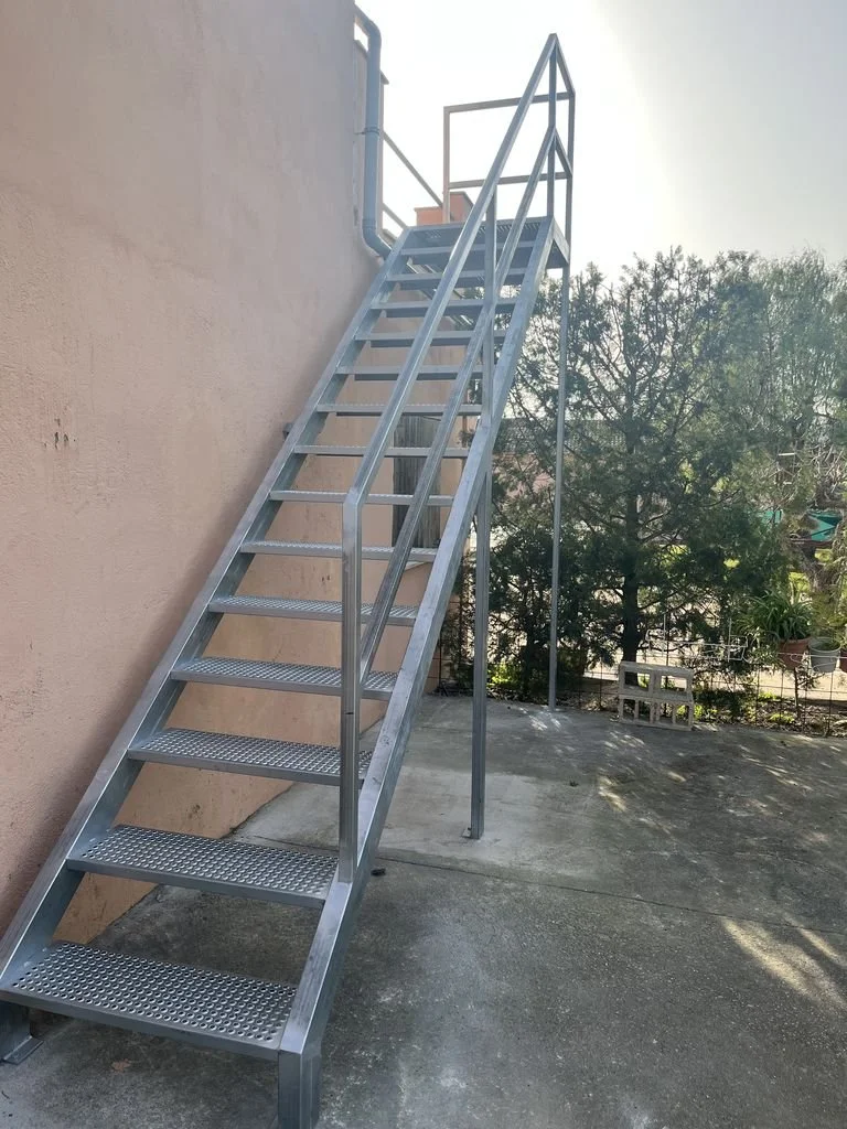 Escalera de metal con pasamanos en un exterior, junto a una pared beige, con árboles y cielo en el fondo.