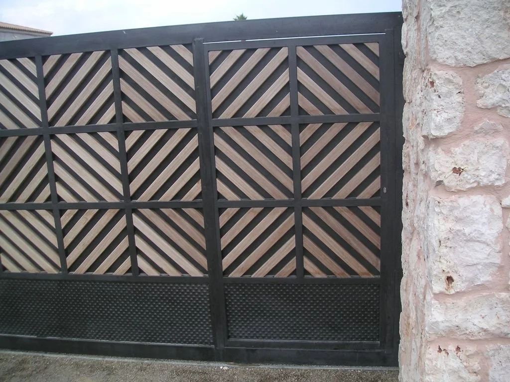 Puerta negra con diseño de listones de madera en patrón diagonal, con parte inferior de malla negra, junto a pared de piedra de color beige.