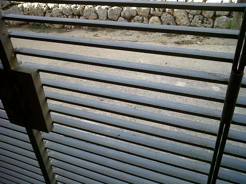 Vista de una reja de metal con barras horizontales, desde el interior de un espacio cubierto, con roca y tierra en el exterior.