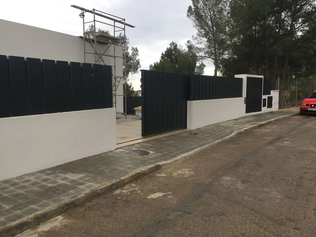 Entrada de casa con muro de bloques blancos y puerta de reja negra, cerca de un árbol y un automóvil rojo en la calle.