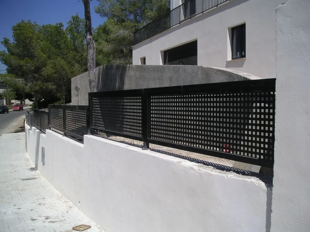Valla negra de metal con cuadrícula en parte superior, en muro blanco frente a calle con árboles y edificios modernos.