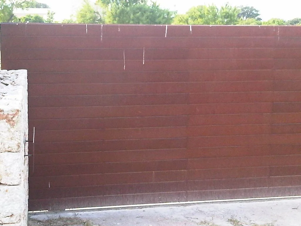 Puerta de hierro de color marrón con estructura de muro de piedra a la izquierda, rodeada de árboles verdes y cielo despejado.