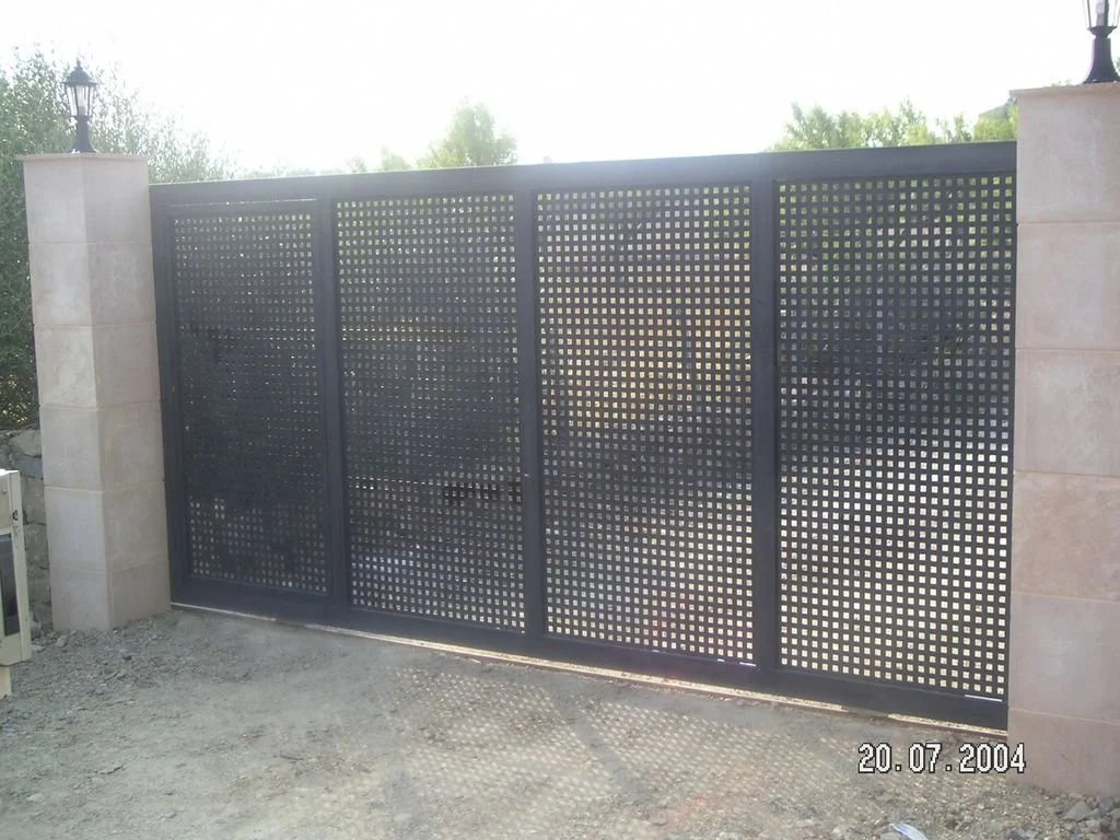 Puerta de metal con paneles perforados en un entorno exterior, flanqueada por pilares de piedra con faroles en la parte superior.