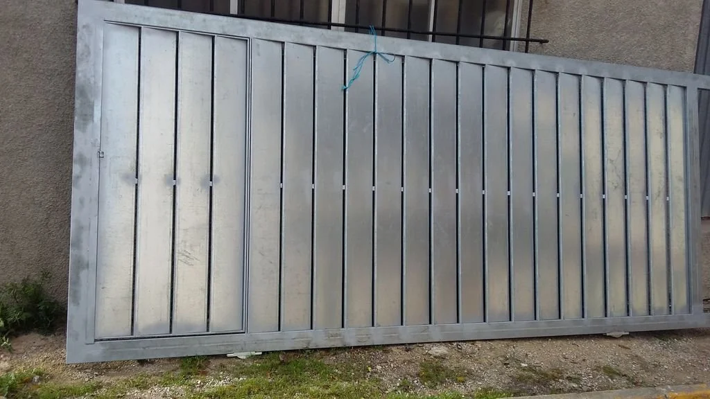 Una puerta metálica grande y plateada apoyada sobre el suelo, en un entorno exterior.