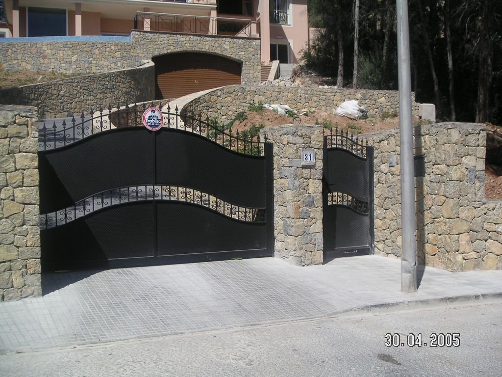 Puerta negra de hierro forjado con muro de piedra en una entrada de casa, en una calle con acera y poste de luz, fecha 30 de abril de 2005