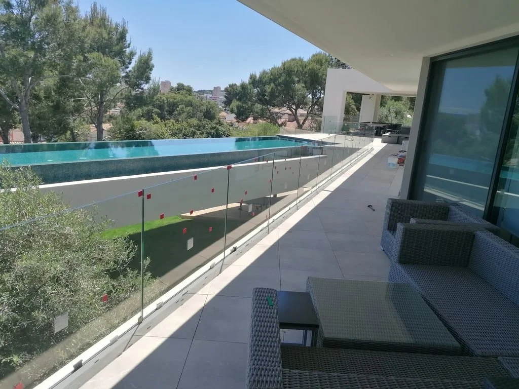 Balcón con sillones de mimbre y mesa de cristal, vista a una alberca y árboles con cielo azul