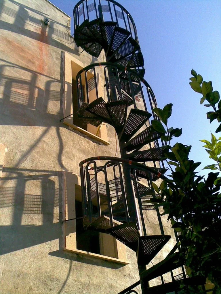 Escalera de caracol de metal negra en el exterior de un edificio de paredes de concreto y una ventana, con sombras proyectadas y un árbol a la derecha, bajo un cielo claro y soleado.