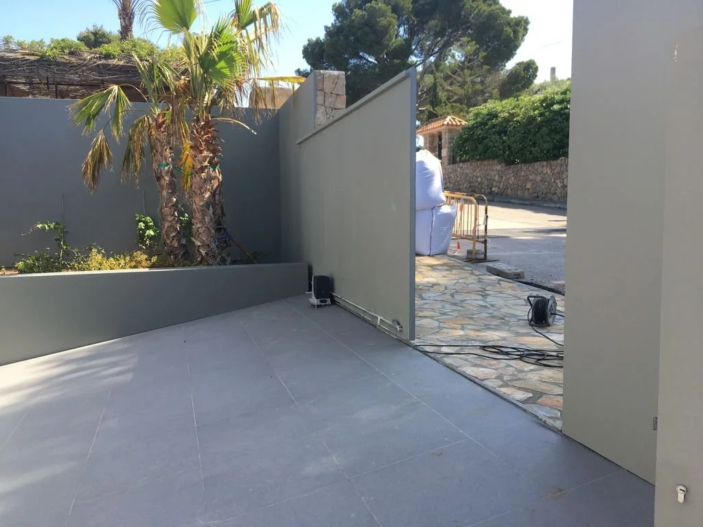 Vista de una pared y puerta de separación en un espacio exterior, con una planta de palma y vegetación en el fondo, y algunas herramientas eléctricas en el suelo.