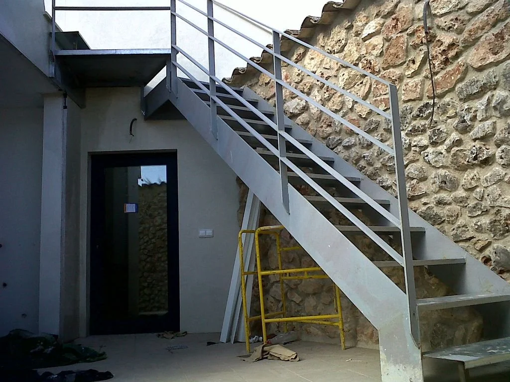 Escalera de metal gris en un patio, junto a una pared de piedra, con una puerta negra y un muro blanco.