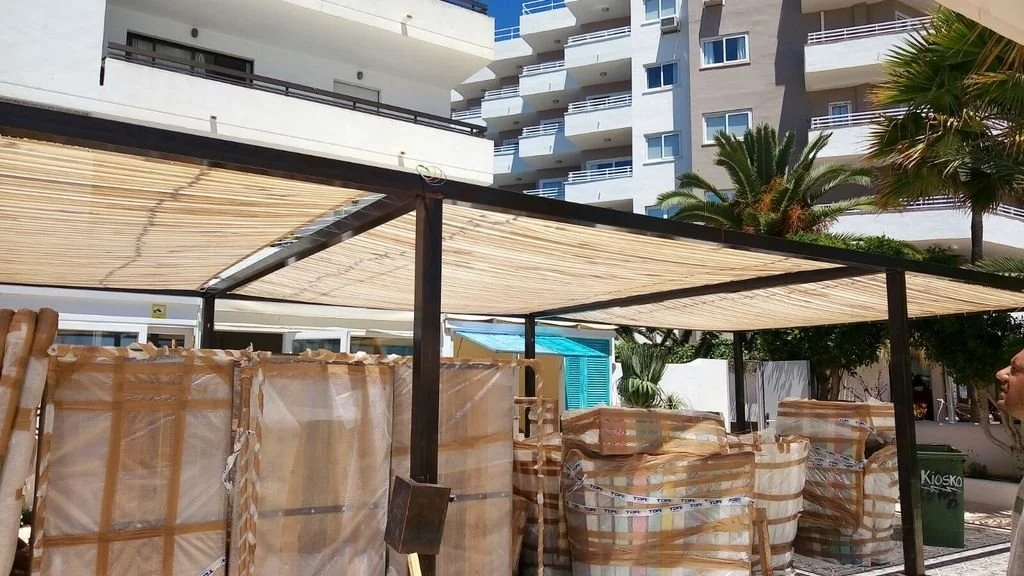 Almacenamiento de sillas apiladas bajo sombra en un área exterior con edificios y palmeras al fondo.