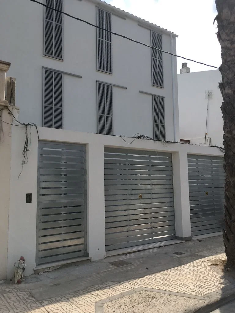 Edificio residencial con fachada blanca y ventanas con persianas grises, reja de metal en la entrada y pared blanca con muro de ladrillo visible en la esquina izquierda.