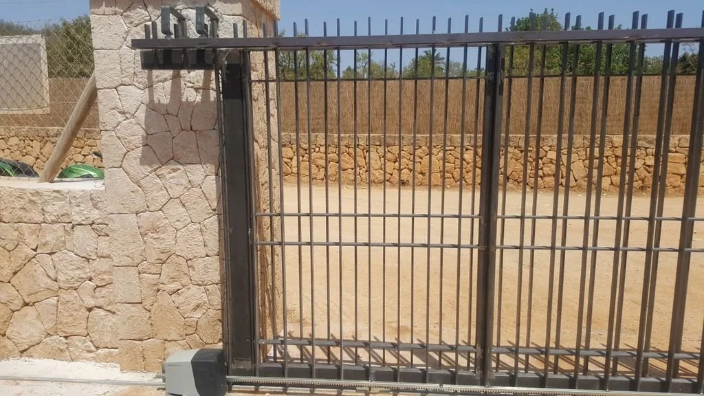 Puerta de rejas de metal con cerradura, en una pared de piedra, con un área abierta de tierra y un fondo de muro de bloques de piedra y vegetación.