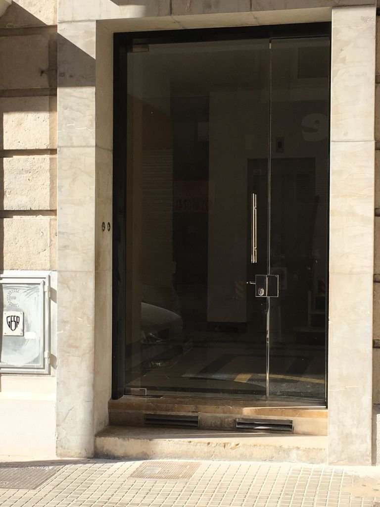 Puerta de vidrio con marco de metal negro en un edificio de piedra, con una pequeña escalinata y rejillas de ventilación en la parte inferior.