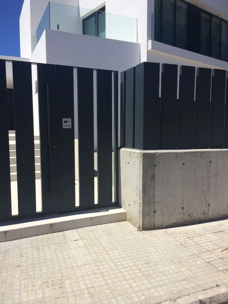 Puerta metálica negra con cerradura en una cerca, en un entorno moderno con edificio de vidrio en el fondo.