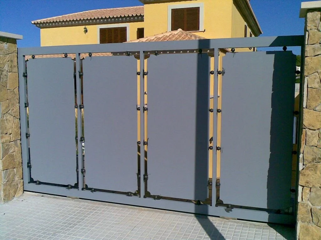 Portón de metal gris con paneles sólidos, instalado entre columnas de piedra, en una vivienda con paredes amarillas y ventanas con persianas de madera.