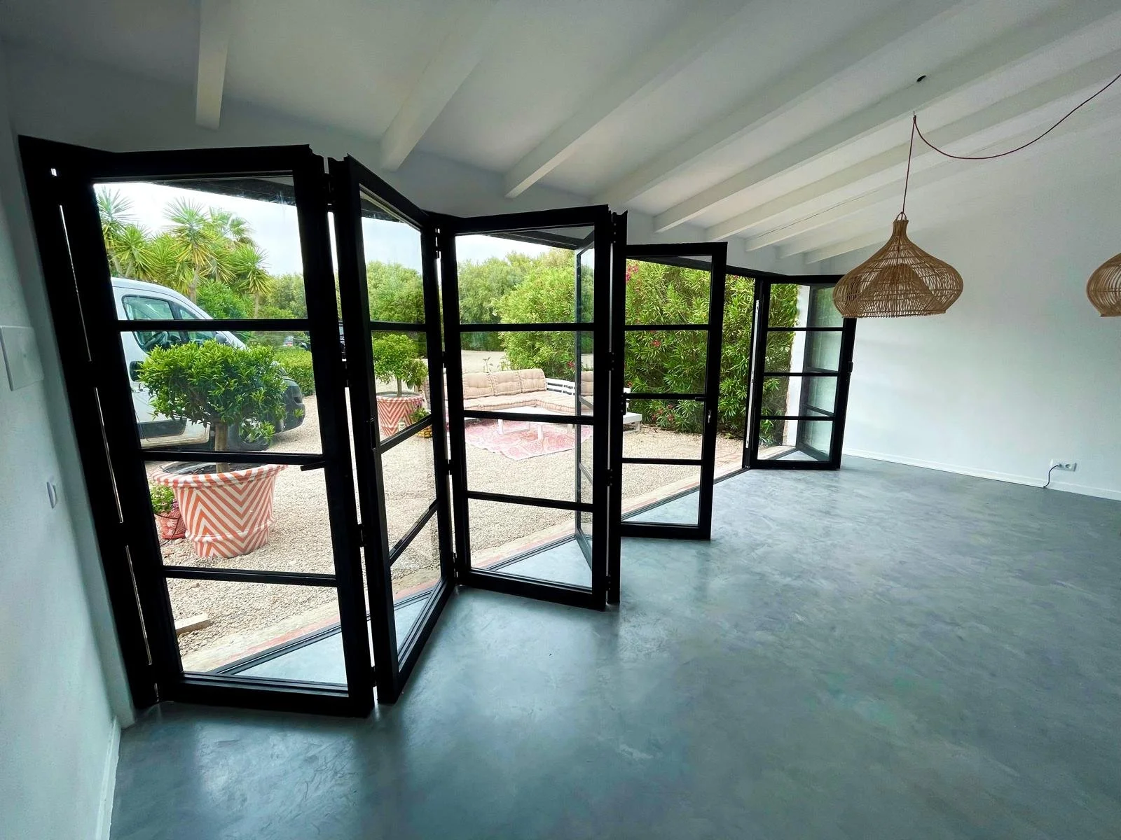 Interior de una sala con ventanas panorámicas abiertas, que dan a un área exterior con plantas y muebles de jardín. Hay lámparas de mimbre colgantes en el techo.