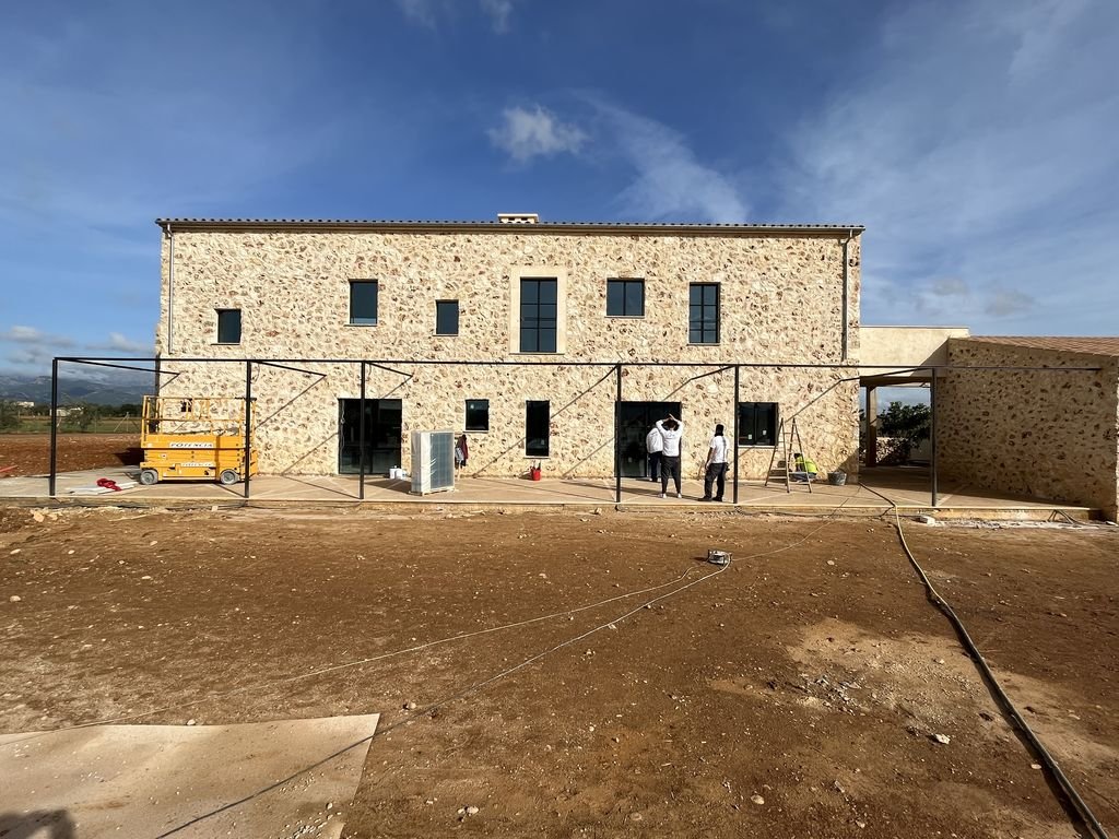 Construcción de un edificio con fachada de piedra y marcos metálicos en el exterior, con varias personas trabajando y herramientas alrededor, en un terreno con tierra y cielo despejado.