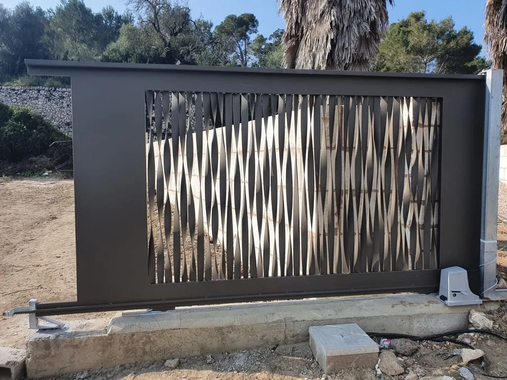 Puerta moderna de cierre eléctrico con diseño de listones en forma de espiral, situada en un entorno exterior con árboles y plantas de fondo.