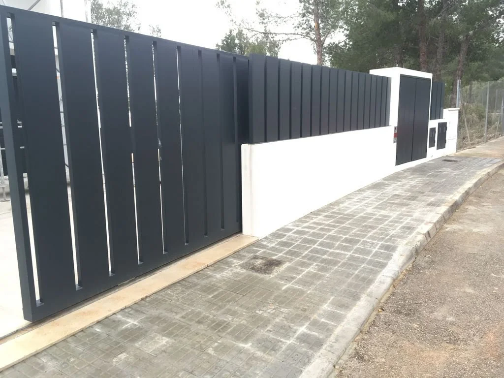 Puerta de reja moderna de metal negro y pared blanca en una acera de pavimento, con árboles y un cerco de alambre en el fondo.