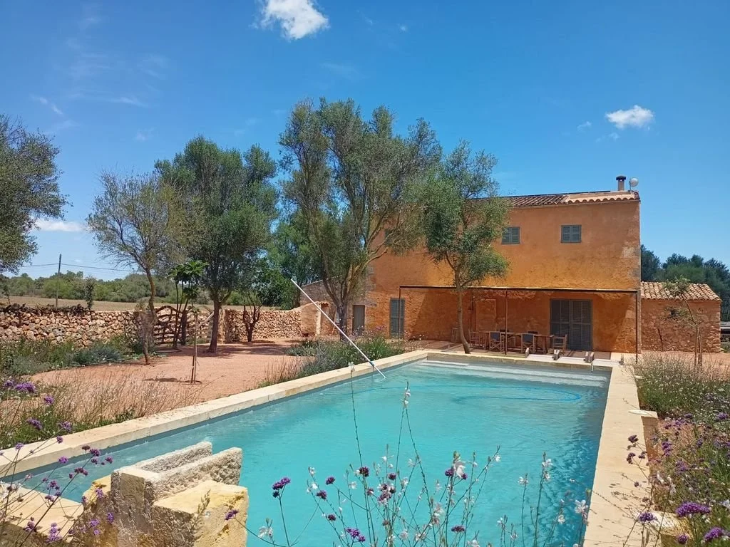 Casa de color terracota con un patio y una piscina en primer plano, rodeada de árboles y vegetación bajo un cielo azul con pocas nubes.