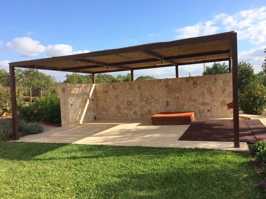 Estructura al aire libre con techo de madera, pared de piedra, suelo de baldosas, área de descanso con cojín y jardín con césped y arbustos.
