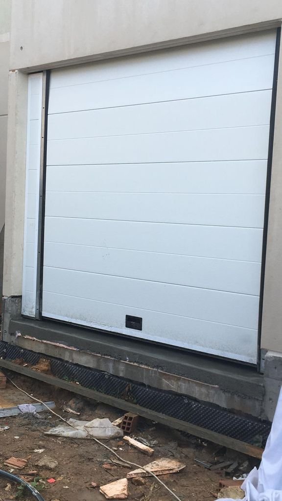Puerta de garaje blanca en construcción con sistema de apertura automática, rodeada de paredes y suelo en obra.