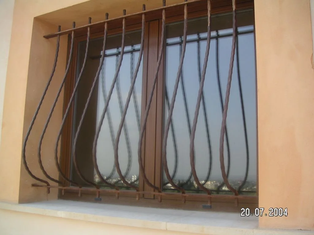 Reja de hierro con barras curvas en una ventana de madera, instalada en un muro de color beige, en un edificio