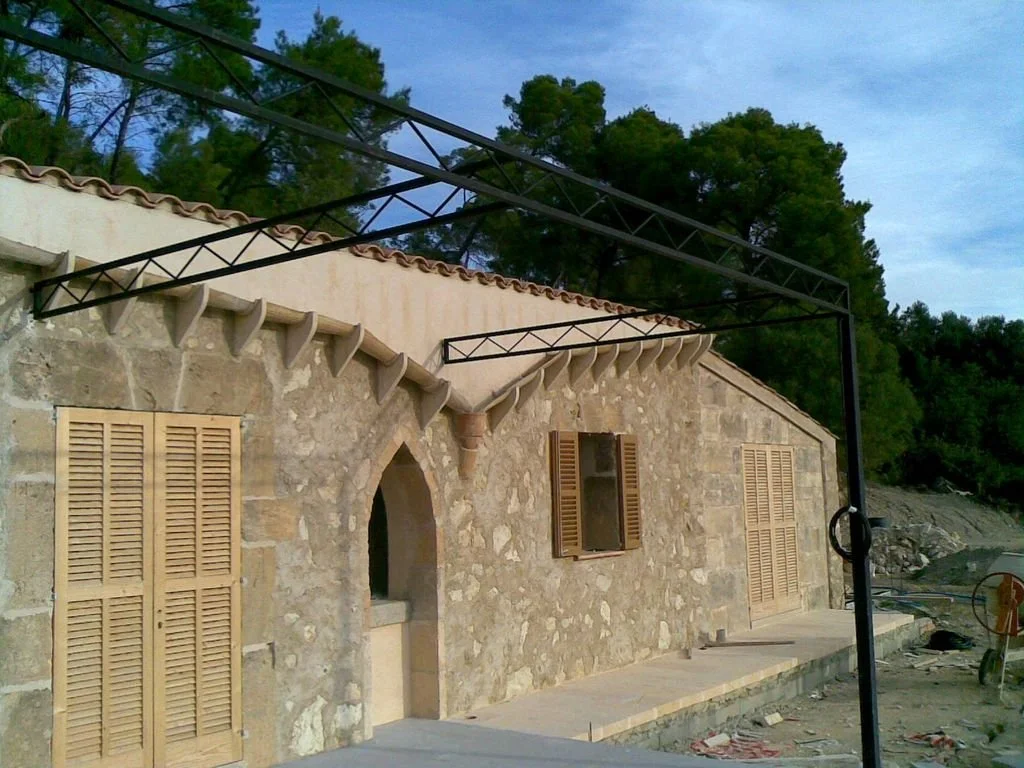 Construcción en progreso en casa de piedra con ventanas de madera y estructura metálica en el techo, en un entorno natural con árboles.
