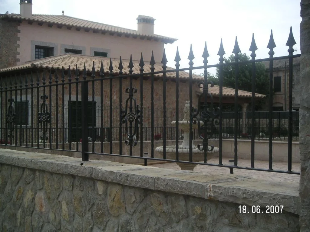 Casa de piedra con ventanas y techo de tejas, cercada por una verja de metal con picos y decoraciones, y una fuente en el patio.