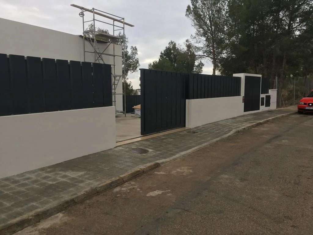 Muro de obra con portón negro y paredes blancas, en proceso de construcción, con escaleras y árboles en el fondo.
