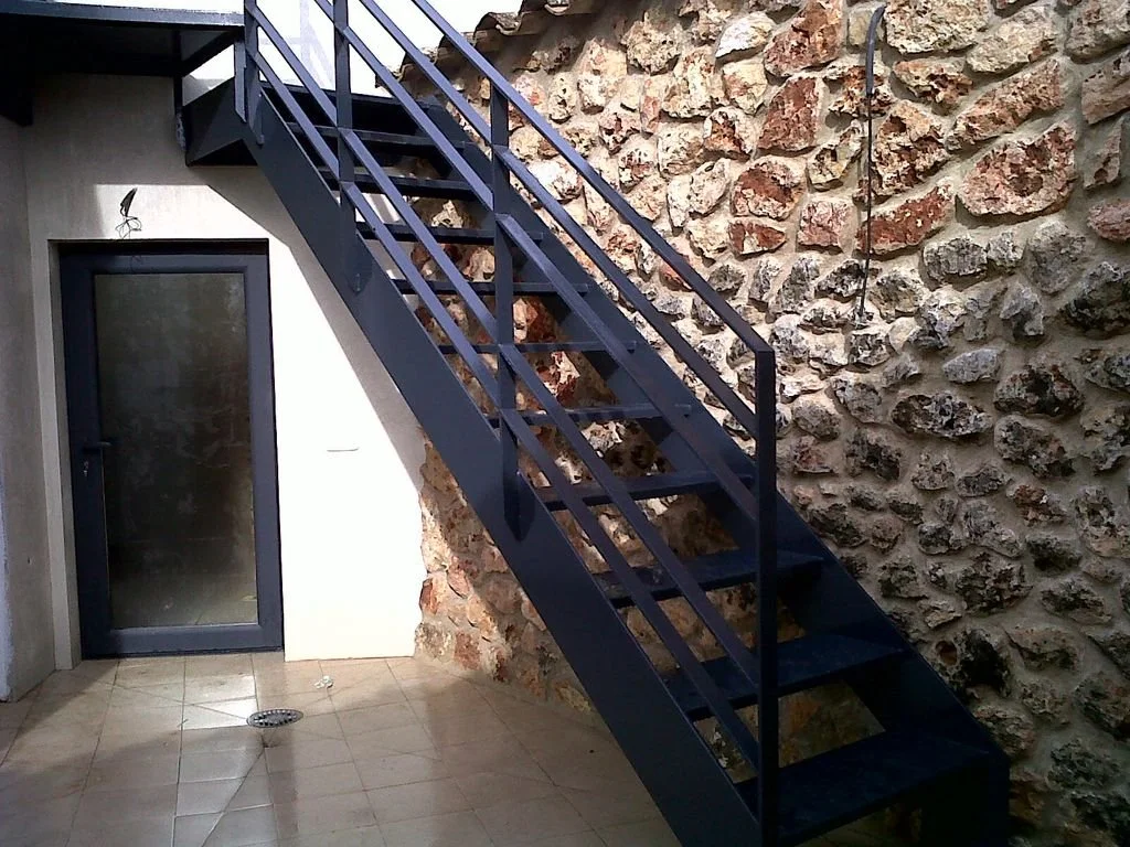 Escalera metálica negra exterior junto a una pared de piedra y una puerta de cristal en un patio con piso de azulejos.