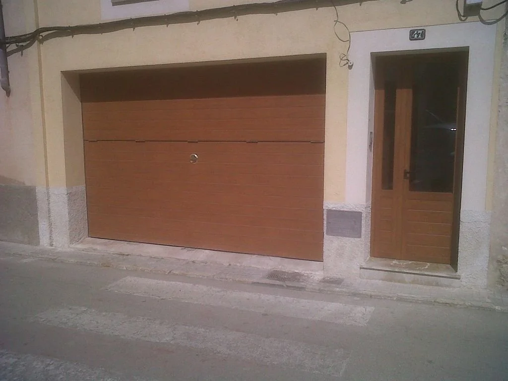Fachada de una casa con portón de cochera de madera y puerta principal de madera con vidrio, en una calle con paso peatonal.