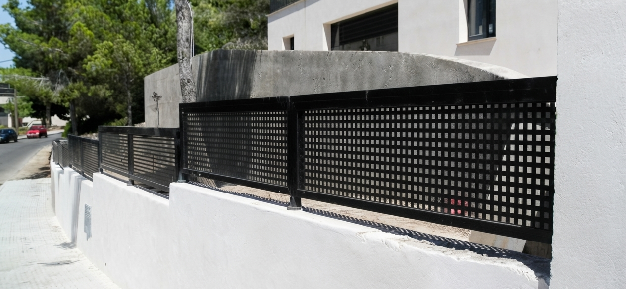 Valla de muro de color negro con diseño de cuadrícula, en un área residencial con calle, árboles y edificios blancos de fondo.