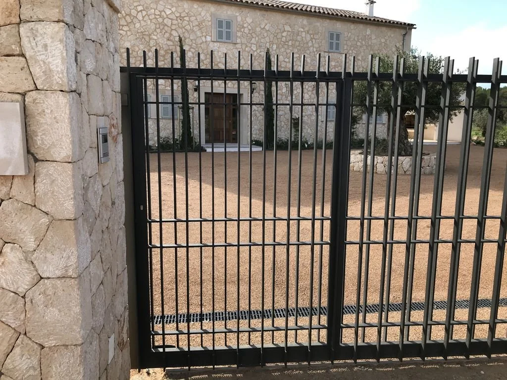 Puerta de reja negra en la entrada de una casa de piedra con un patio de tierra.