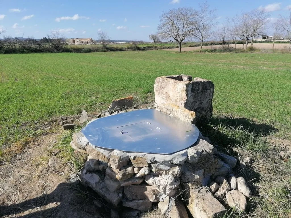 Pozo de agua rústico en campo abierto con cielo despejado y árboles sin hojas en el fondo